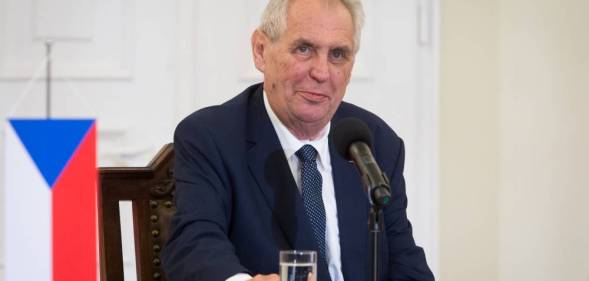Czech president Miloš Zeman sits at a table in front of a microphone and glass of water while wearing a white button up shirt, blue tie and dark blue suit jacket