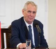 Czech president Miloš Zeman sits at a table in front of a microphone and glass of water while wearing a white button up shirt, blue tie and dark blue suit jacket