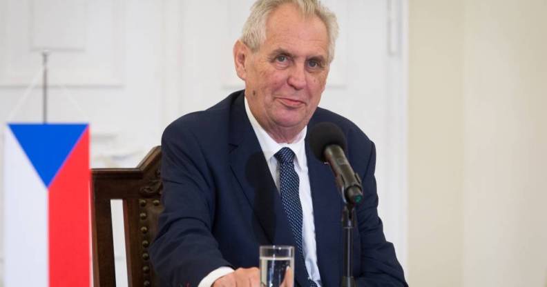 Czech president Miloš Zeman sits at a table in front of a microphone and glass of water while wearing a white button up shirt, blue tie and dark blue suit jacket