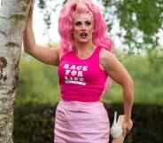 Drag Race UK star Ella Vaday wears a pink wig, hot pink sleeveless shirt with a white 'Race for Life' logo on it and baby pink short skirt. She poses with one hand on a tree while holding one foot, which has a white high heel on it, in her other hand