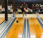 Michelle Guzowski throws a bowling ball down the lane of a bowling alley while other people in the background watch her bowl