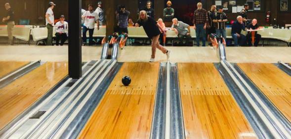 Michelle Guzowski throws a bowling ball down the lane of a bowling alley while other people in the background watch her bowl