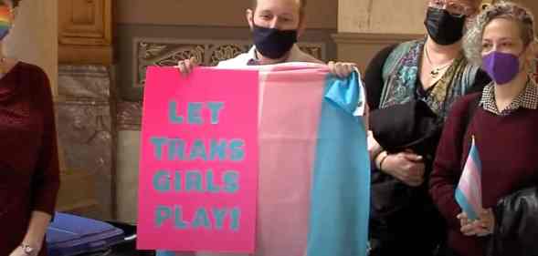 A person wearing a black face mask holds up a trans Pride flag and a sign reading "Let trans girls play" in protest of trans sports bans being passed