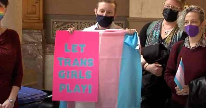 A person wearing a black face mask holds up a trans Pride flag and a sign reading "Let trans girls play" in protest of trans sports bans being passed