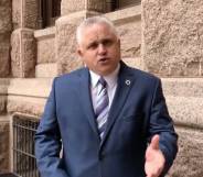 Republican Texas lawmaker Bryan Slaton stands in front of a brick building while wearing a white button up shirt, blue striped tie and blue suit jacket. He is gesturing with one hand towards the camera