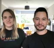 Hannah and Jake Graf sit in their house while wearing black shirts. Hannah wears a shirt that says 'we are one' in rainbow font while Jake sits next to her also wearing a black shirt. In the background, there are pillows and blankets on a yellow chair