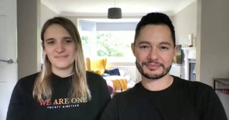 Hannah and Jake Graf sit in their house while wearing black shirts. Hannah wears a shirt that says 'we are one' in rainbow font while Jake sits next to her also wearing a black shirt. In the background, there are pillows and blankets on a yellow chair