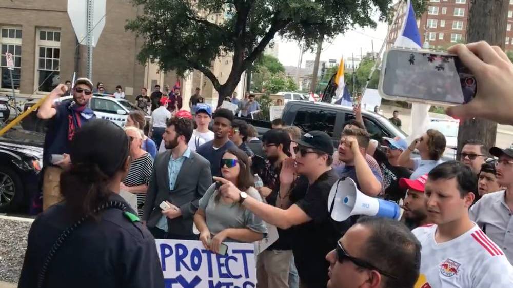 Protestors gather outside a bar in Texas with one person shouting into a megaphone while another person holds a sign reading 'Protect TX kids'