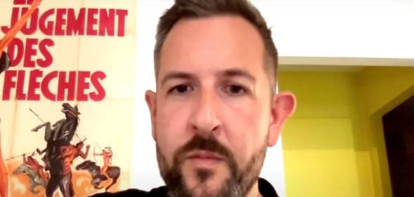 James McFadzean sits in a room wearing a dark shirt. The wall behind him is white and there is a poster with a person rising a bucking horse with words in French written on it
