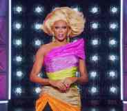 RuPaul wears a one should dress in pink, yellow, tan and orange with a blonde wig on the set of Drag Race All Stars 7