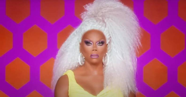 RuPaul wears a white blonde wig and yellow dress as the drag queen sits in front of a pink and orange patterned background