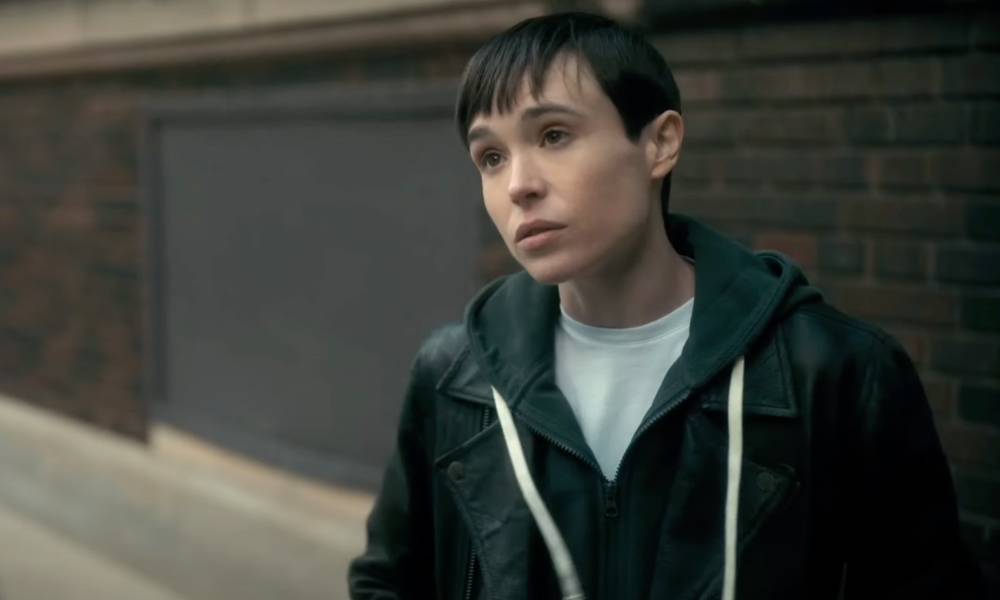 Elliot Page appears as his character from the Netflix show The Umbrella Academy. The character, Viktor, is wearing a white t-shirt and blue hooded sweatshirt as he stands outside and talks to someone off screen
