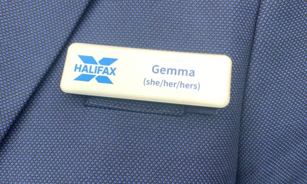 A Halifax employee wears a blue blazer and name badge which includes the name 'Gemma' and her pronouns 'she/her/hers' as well as the blue Halifax logo