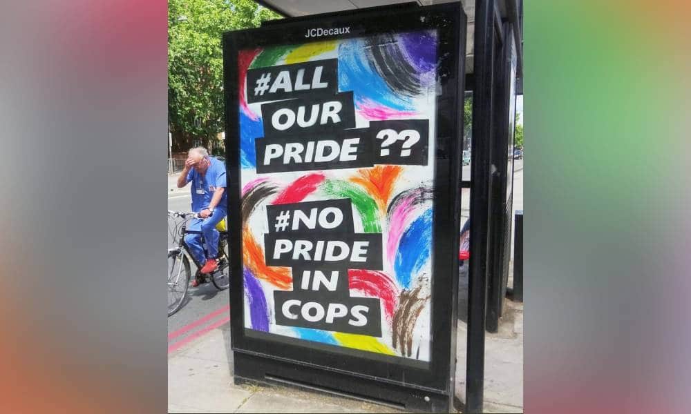 A colourful sign reading 'All Our Pride?? No Pride in Cops' is displayed outside a bus stop in London