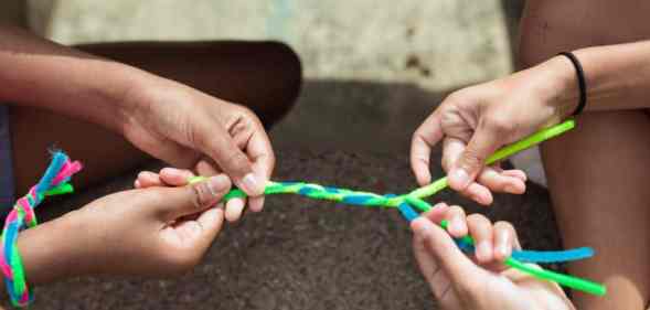 Two people hold fuzzy pipe cleaners which are coloured blue and green which they are braiding together