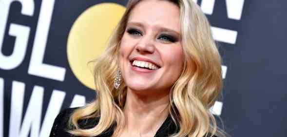 Kate McKinnon smiles as she wears a black outfit and her blonde hair is styled in waves. She is standing in front of a billboard for the the 77th Annual Golden Globe Awards