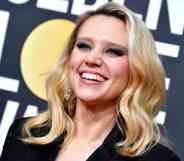 Kate McKinnon smiles as she wears a black outfit and her blonde hair is styled in waves. She is standing in front of a billboard for the the 77th Annual Golden Globe Awards