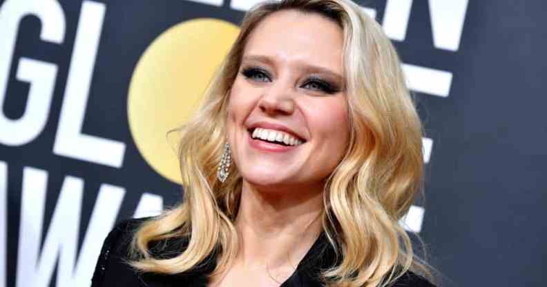 Kate McKinnon smiles as she wears a black outfit and her blonde hair is styled in waves. She is standing in front of a billboard for the the 77th Annual Golden Globe Awards