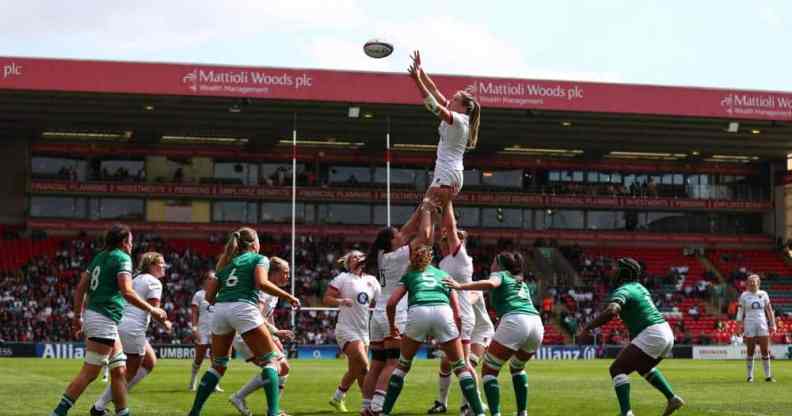 Several women wearing green or white rugby uniforms surround one player who is being held up by their teammates to catch the ball which is flying in the air towards them