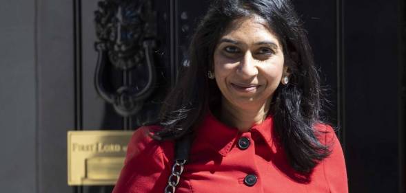 Suella Braverman wears a red coat with black buttons as she smiles to camera while standing in front of a black door