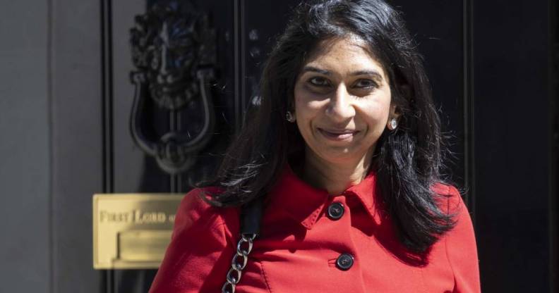 Suella Braverman wears a red coat with black buttons as she smiles to camera while standing in front of a black door