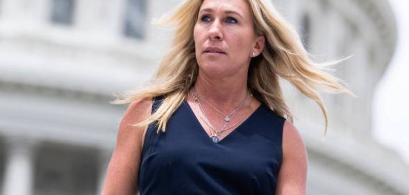 Marjorie Taylor Greene wears a dark blue dress as she stares off camera with her blonde hair blowing in the background. There is a white building seen in the background