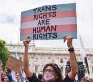 A person wearing a face mask designed in the colours of the trans Pride flag holds up a sign with the colours of the trans Pride flag that reads "Trans rights are human rights" above their head during a protest