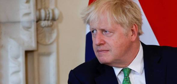 Boris Johnson wears a white button up shirt, green tie with a black shirt as he sits down and stares at someone off camera. There is a blue, red and white flag in the background