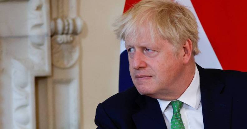 Boris Johnson wears a white button up shirt, green tie with a black shirt as he sits down and stares at someone off camera. There is a blue, red and white flag in the background