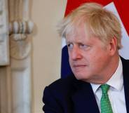 Boris Johnson wears a white button up shirt, green tie with a black shirt as he sits down and stares at someone off camera. There is a blue, red and white flag in the background