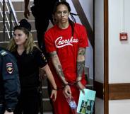 Brittney Griner wears a red short sleeved t-shirt with white writing on it as she is led down a stairway in Russia in handcuffs