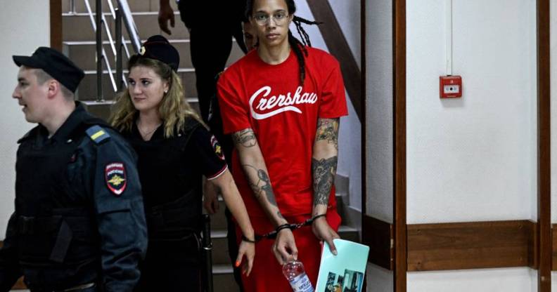 Brittney Griner wears a red short sleeved t-shirt with white writing on it as she is led down a stairway in Russia in handcuffs