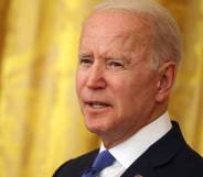 President Joe Biden is seen wearing a white button up shirt, blue tie and dark suit jacket as he stands in front of a yellow background