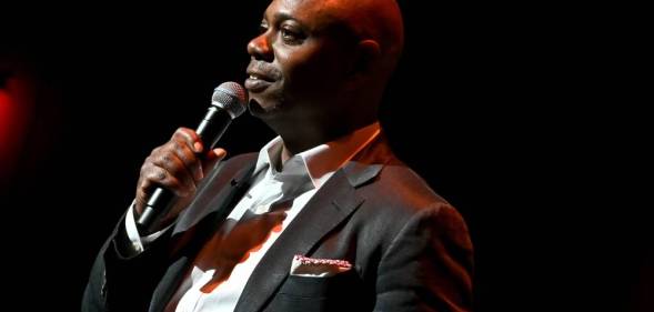 Dave Chappelle wears a white button up shirt, dark suit jacket and has something sticking out of the jacket's breast pocket as he speaks into a microphone he is holding is his hand