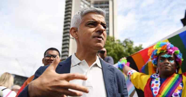 Major of London Sadiq Khan wears a white button up shirt and grey jacket as he attends Pride in London celebrations. A person in the background wears rainbow clothing as they hold up an LGBTQ+ Pride flag