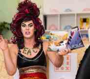 Aida H Dee, a drag queen and founder of Drag Queen Story Hour UK, wears a black top and rainbow bottoms as she reads from a book in front of a group of children. She is gesturing with both her hands in the air like an animal roaring