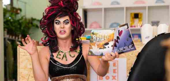 Aida H Dee, a drag queen and founder of Drag Queen Story Hour UK, wears a black top and rainbow bottoms as she reads from a book in front of a group of children. She is gesturing with both her hands in the air like an animal roaring