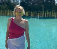Rebel Wilson wears a pink off the shoulder swimsuit with a white cloth around her hips as she stands near a pool