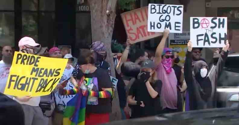Several people gather outside of a drag event in Houston, Texas. One person holds up a white sign reading 'Bigots go home', another sign reads 'Hail Satan', a yellow sign reads 'Pride means fight back' and one more sign reads 'Bash the fash'