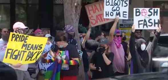 Several people gather outside of a drag event in Houston, Texas. One person holds up a white sign reading 'Bigots go home', another sign reads 'Hail Satan', a yellow sign reads 'Pride means fight back' and one more sign reads 'Bash the fash'