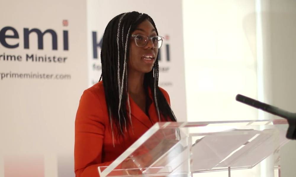 Kemi Badenoch stands at a transparent podium as she speaks to a crowd gathered off screen. She is wearing a red-orange dress with her hair styled in braids with white signs seen in the background.