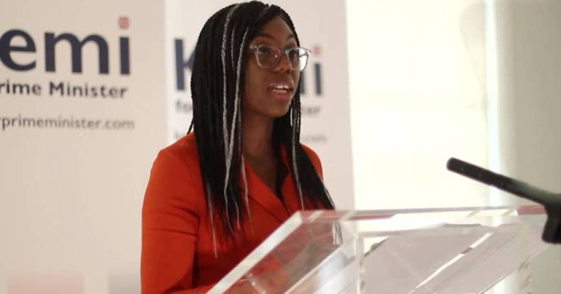 Kemi Badenoch stands at a transparent podium as she speaks to a crowd gathered off screen. She is wearing a red-orange dress with her hair styled in braids with white signs seen in the background.