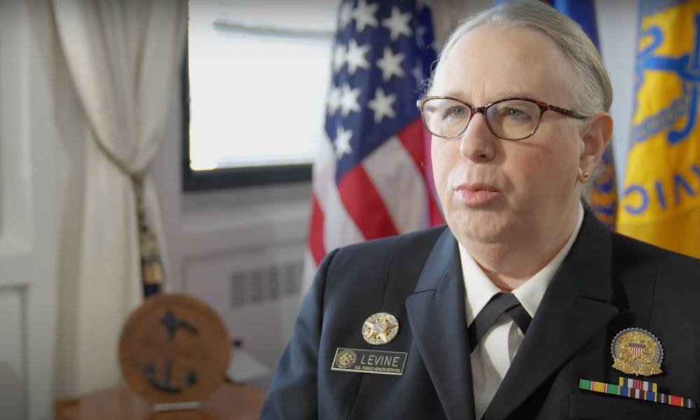 Dr Rachel Levine speaks to someone off camera while she is wearing a white shirt, dark jacket and pins denoting her status as a four-star admiral. There is a US flag seen in the background