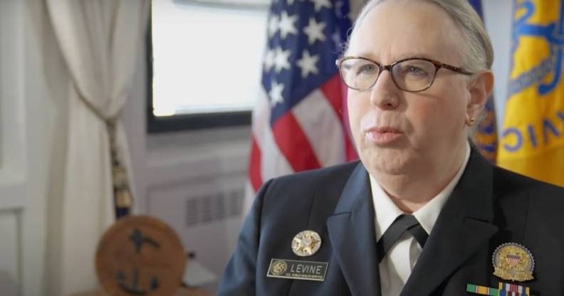 Dr Rachel Levine speaks to someone off camera while she is wearing a white shirt, dark jacket and pins denoting her status as a four-star admiral. There is a US flag seen in the background