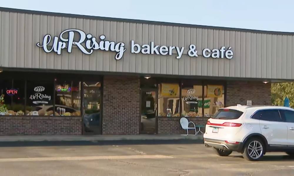 The image shows a business front with the words 'UpRising Bakery and Cafe' written in white. A white car is parked outside in the parking lot