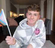 Adam Beales, known as Adam B on YouTube, wears a grey and white checkered fuzzy sweatshirt with a pink smiley face on the chest. He is waving a progressive Pride flag in one hand.