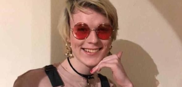 Alice Litman with short blonde hair, wearing sunglasses, black dungarees and a pink t-shirt, smiling as the sun shines on her