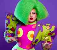 Drag Race UK star Choriza May wears a green wig, colourful outfit with large shoulder pads as she stands in front of a purple background