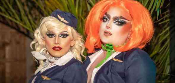Two drag performers stand side by side, one with blonde hair and another with orange hair, as they appear to wear what looks like air cabin crew uniforms