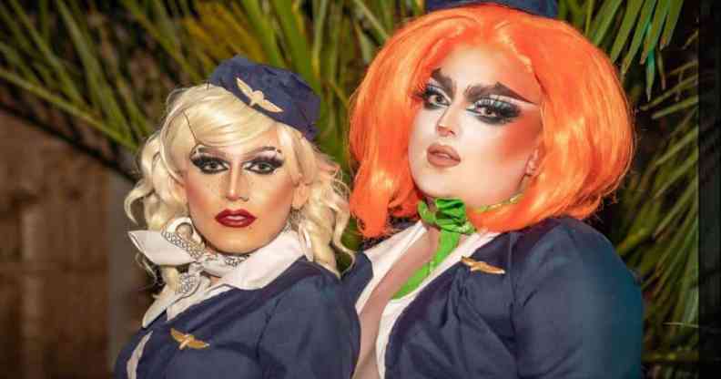 Two drag performers stand side by side, one with blonde hair and another with orange hair, as they appear to wear what looks like air cabin crew uniforms